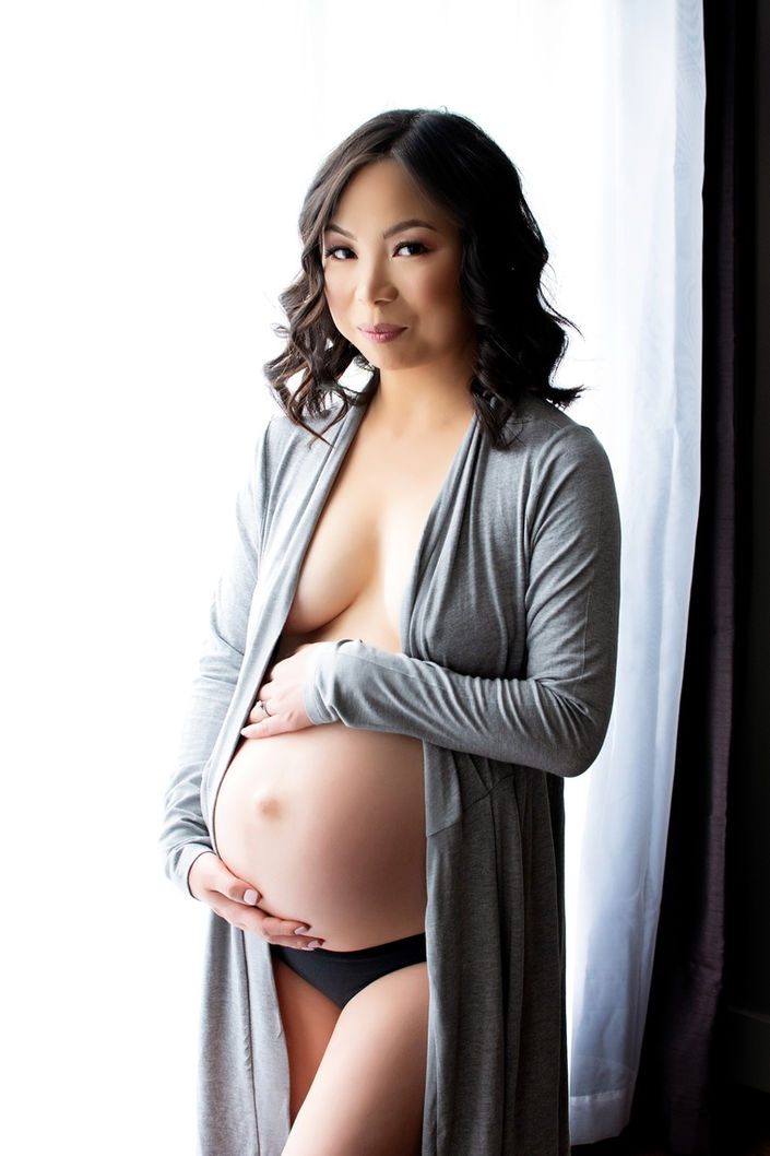 Pregnant woman wearing an open robe and lingerie, gently holding her belly in a maternity boudoir photoshoot