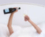 close up of arms holding a champagne bottle and glass under white sheets on a white bed.