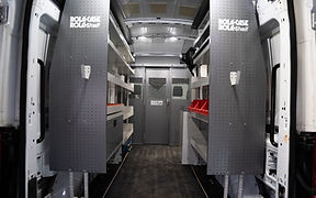 Interior of an upfitted cargo van with gray shelving, pegboard panels, storage bins, and a metal partition separating the cargo area from the cab.