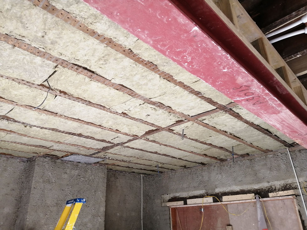 Construction site interior with insulated ceiling panels, exposed rebar, a red beam, hanging wires, and a yellow ladder below