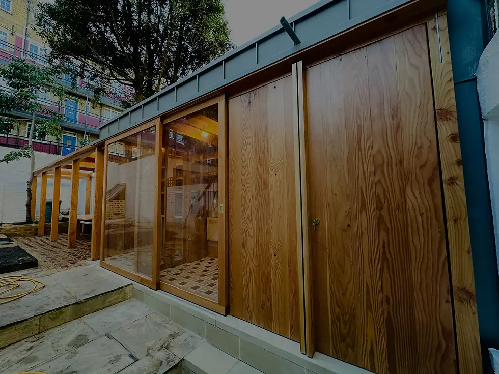 Wooden structure with large glass doors, set in a paved yard with trees. A nearby building has colorful balconies. Evening lighting creates a warm mood.