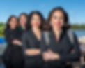 Four smiling professional women wearing black blazers, arms crossed outdoors.