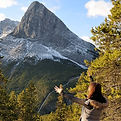 View of a mountain, fall folliage at the base with someone at the base with arms extended reaching out (Canmore, Alberta)