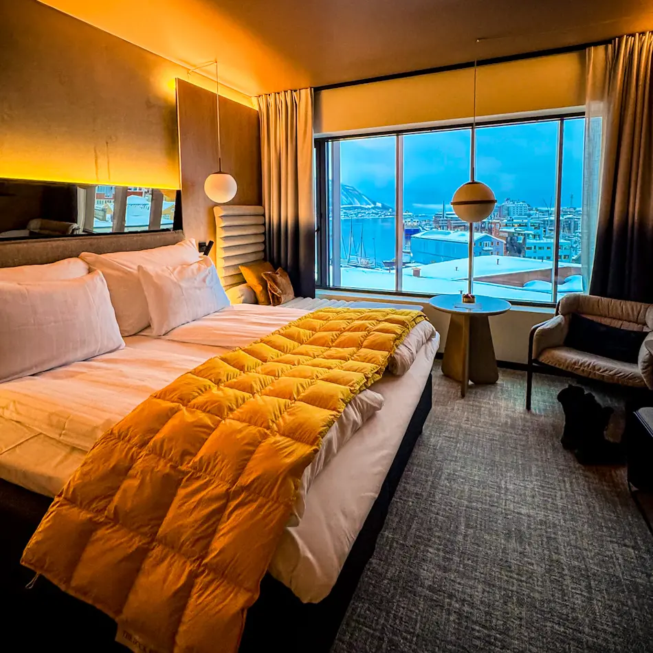 Standard King Room at The Dock 69°39 by Scandic, one of the luxury hotels in Tromso, with mustard quilt, large window overlooking harbour and snowy mountains.