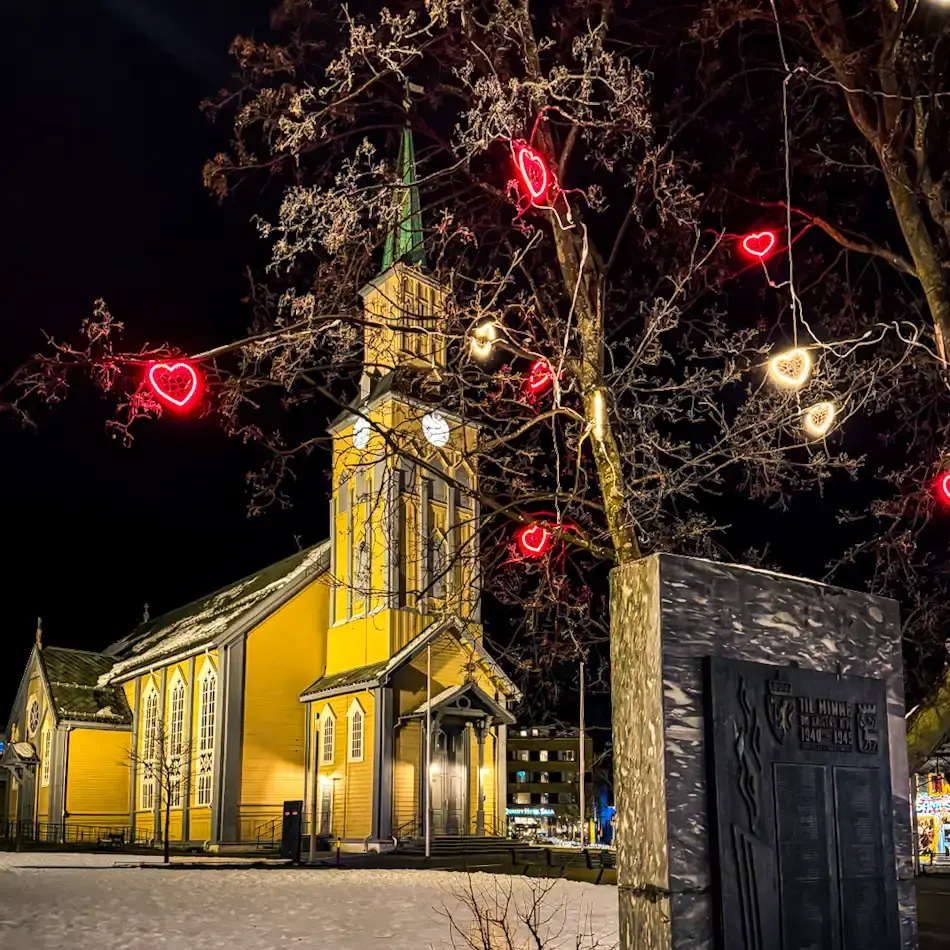 Tromsø Cathedral at night illuminated in warm yellow light with festive heart-shaped decorations in the trees and snow on the ground, highlighting historic things to do in Tromso in winter.