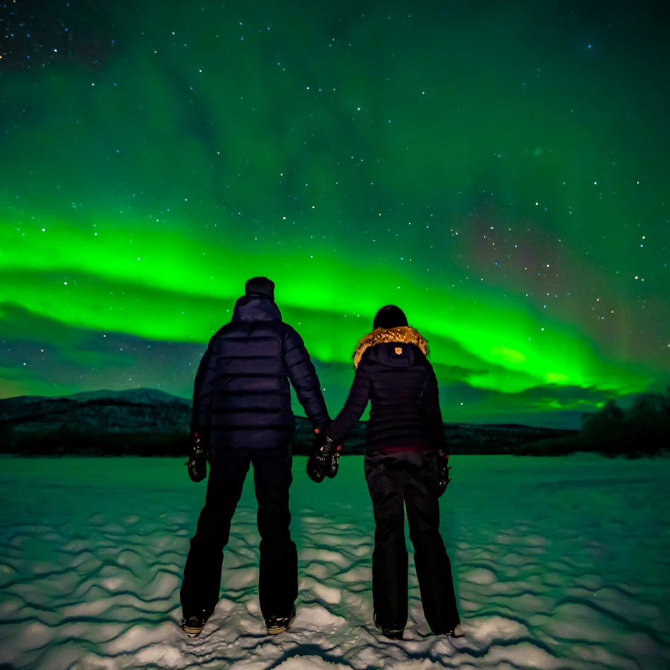 James & Shal standing beneath bright green northern lights on a aurora chase during our Tromso itinerary.