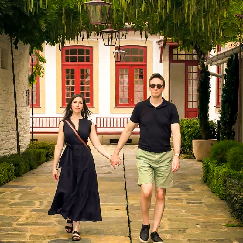 Shal and James holding hands walking down a path with a colonial looking building in the background and an arch of hanging vines and lanterns above their heads.