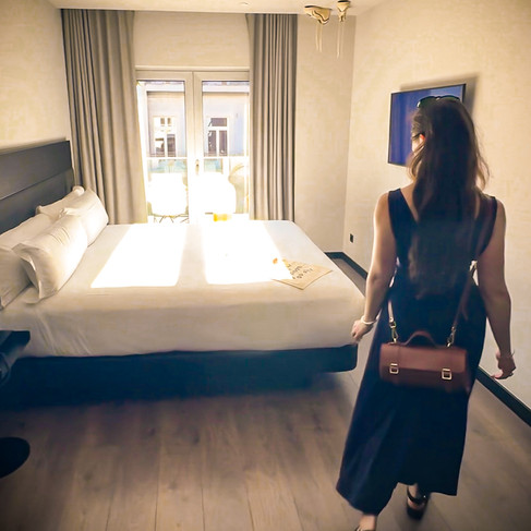 Shal walking into a minimally decorated hotel room with a bed with crisp white lines and sneakers walking on the ceiling.