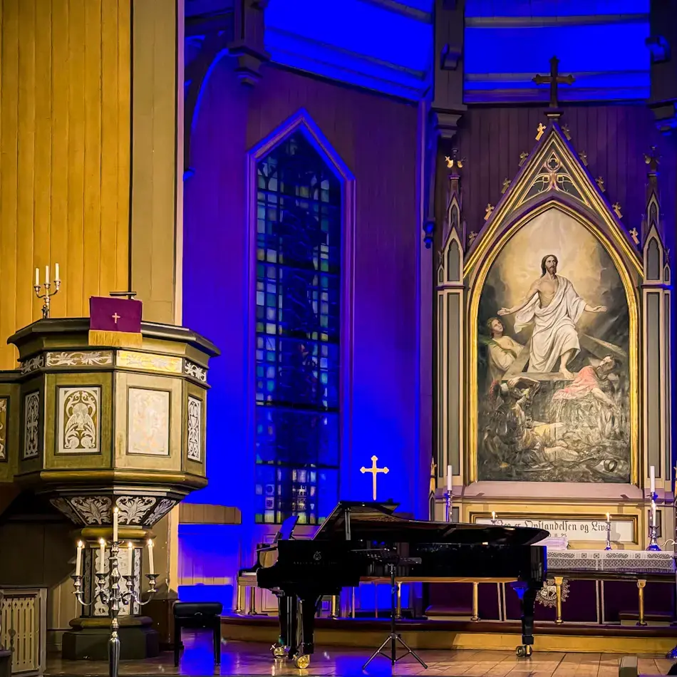 Interior of Tromsø Cathedral lit in blue and gold during a midnight concert, with a grand piano in front of the altar and stained glass windows behind, showcasing cultural things to do in Tromso.
