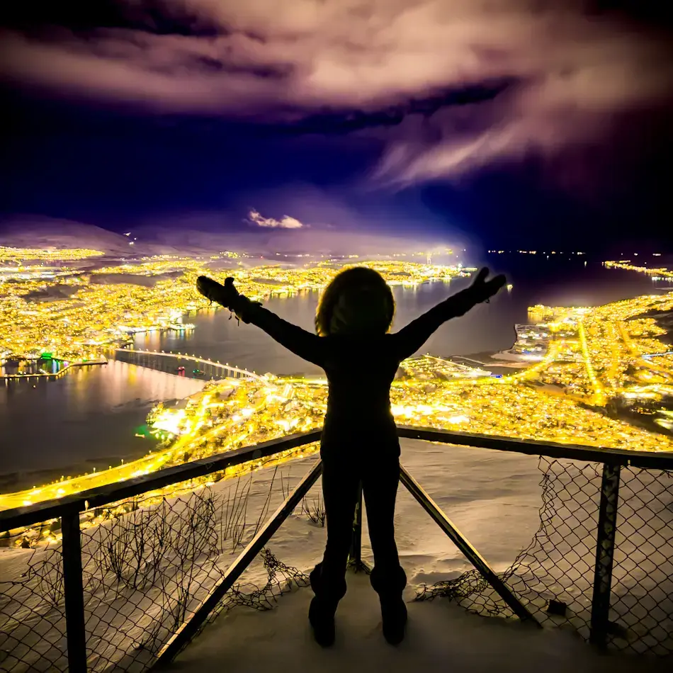 Shal in silhouette standing at the Fjellheisen viewpoint above Tromsø at night with arms raised, overlooking the glowing city lights, harbour and Tromsø Bridge below, capturing panoramic things to do in Tromso.