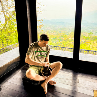 James sitting cross-legged on a wooden floor, playing singing bowls beside large windows with views of rice fields in Vietnam.