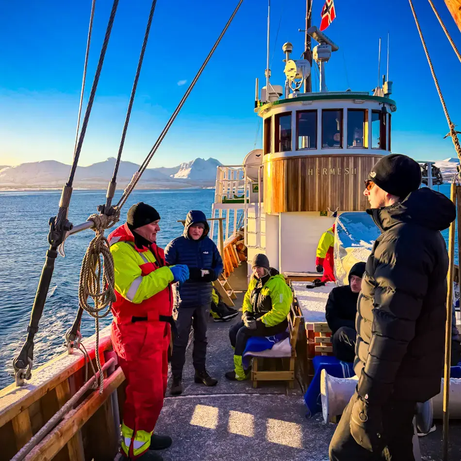 Small group of travellers standing on the deck of a wooden boat in the Arctic during winter, listening to a guide in red overalls while mountains and blue water surround them. A scenic example of things to do in Tromso.