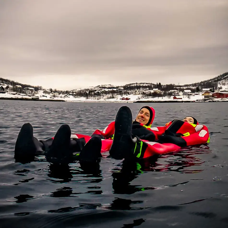 James and Shal lying back and drifting on icy fjord water during Arctic floating, wearing bright red survival suits with snowy Norwegian hills in the background.