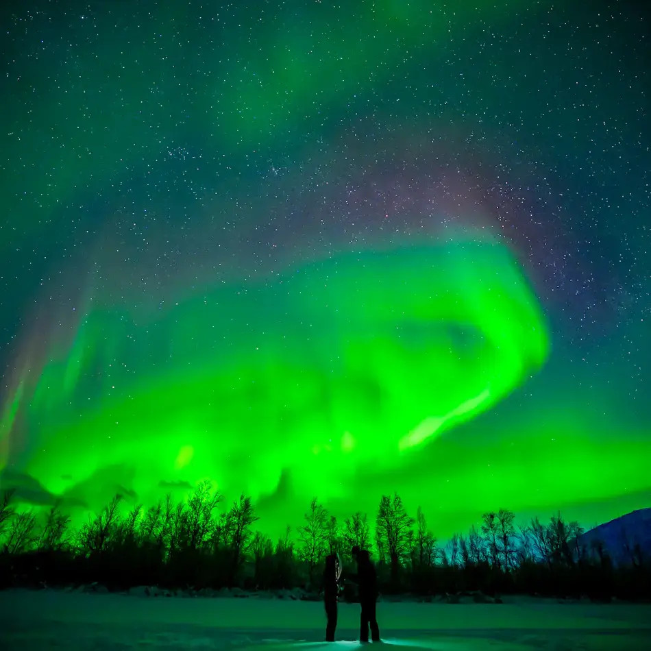 Shal and James standing on snowy Arctic landscape beneath vivid green aurora borealis and star-filled sky in Finland during a Tromso northern lights tour.