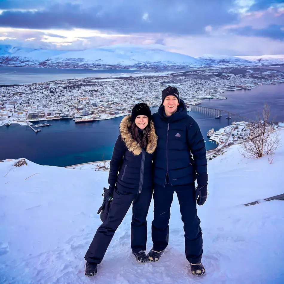 Shal and James standing in the snow at the Fjellheisen viewpoint above Tromsø during winter daylight, overlooking the harbour, Tromsø Bridge and surrounding snowy mountains, highlighting scenic things to do in Tromso.
