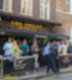 Outside The Toucan pub with many customers chatting and drinking Guinness. The bar has standout black and amber décor that matches the colour of a Toucan. Two security guards man the door.