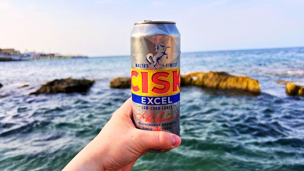A silver can of Cisk Excel beer held in front of the Mediterranean Sea at sunset, with rocks visible in the background.