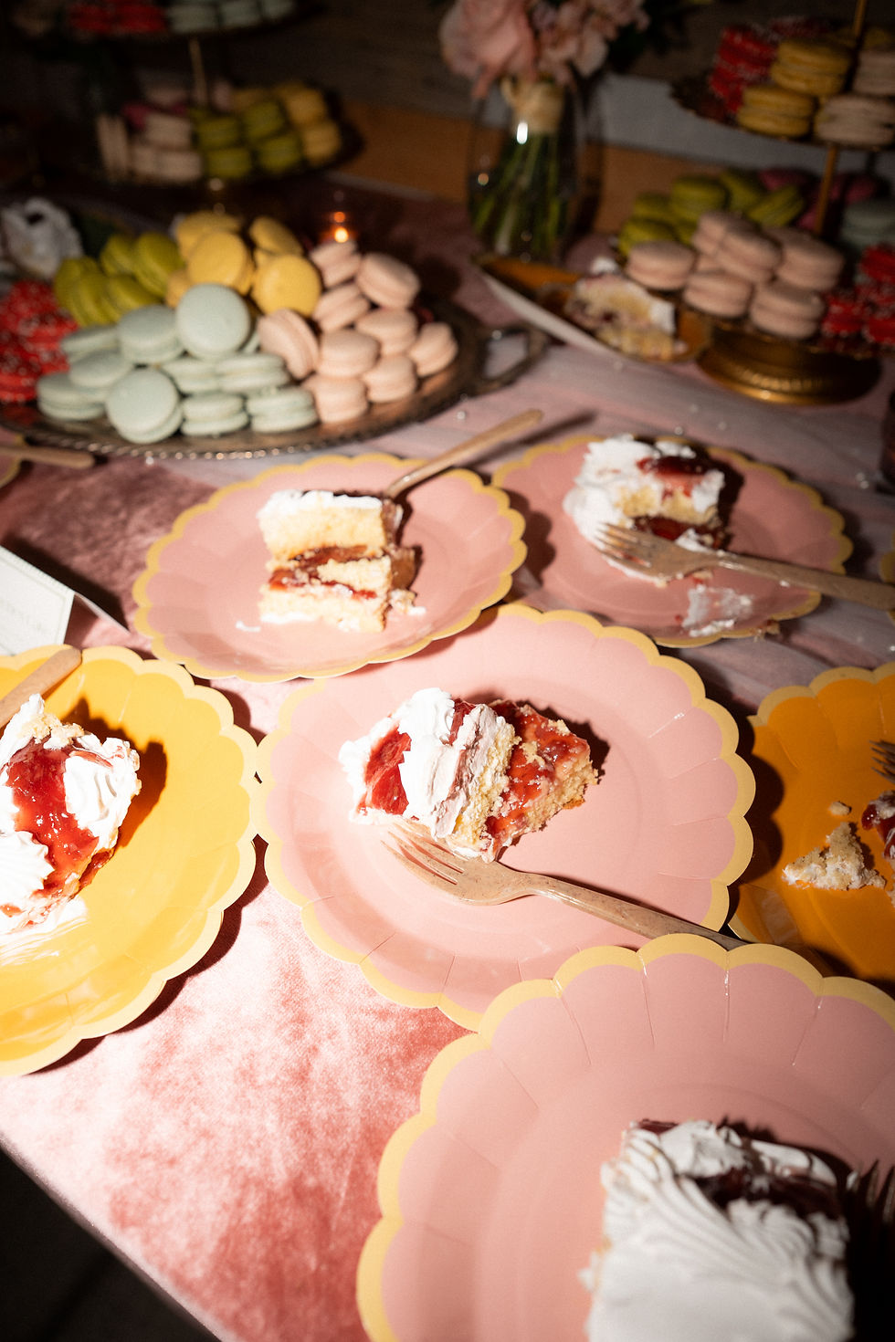 Slices of cake on pink and yellow scalloped plates with wooden forks. Background features stacked macarons in various colors.
