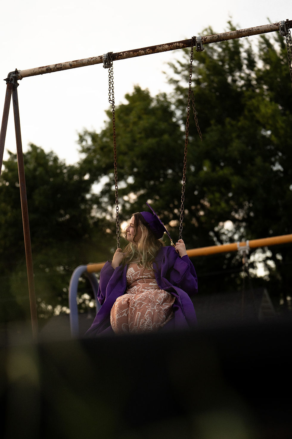A person in a purple graduation gown swings joyfully at a playground, with green trees in the background under a bright sky.