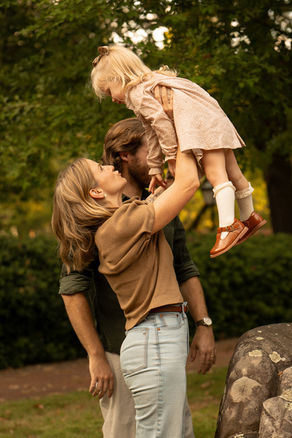 Woman lifts smiling child in park, man stands behind. Lush green trees and rocks create a warm, happy outdoor setting. Photos by Arkansas Photographer, Kalen Kemp
