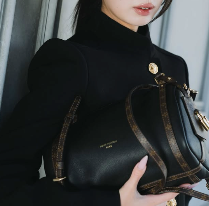 Person in black coat holding a black Louis Vuitton L'Express bag with gold accents. Background is a simple, neutral wall. Mood is stylish and elegant.