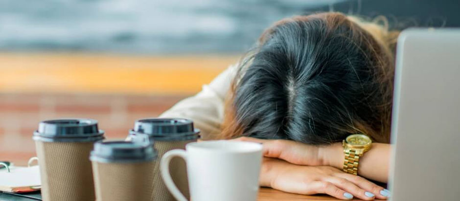 A woman with her head on her hands at her desk, with 4 cups of coffee, clearly exhausted.