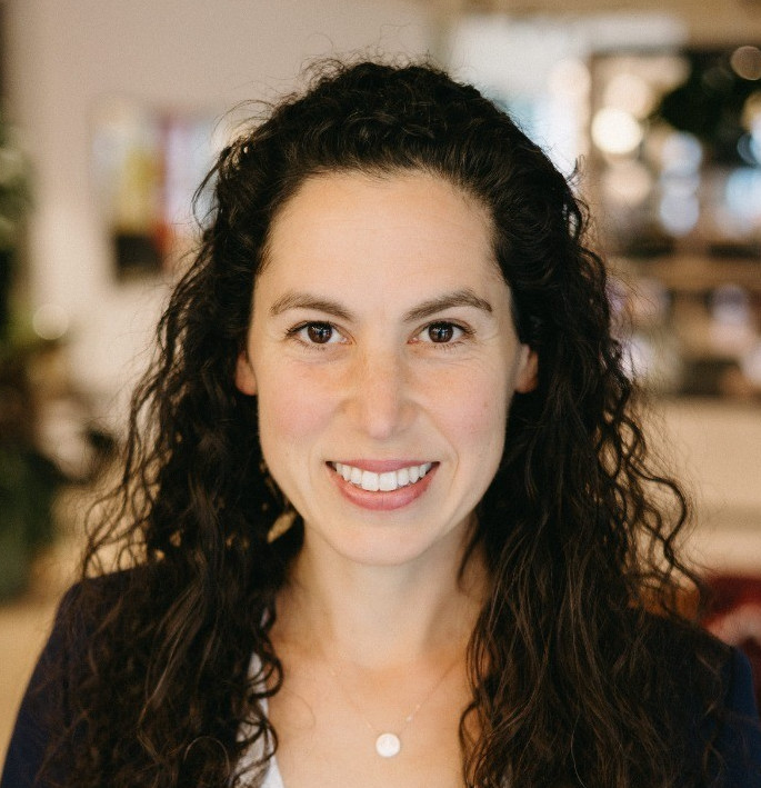 Square portrait headshot of a person with long dark curly hair and light skin, smiling, in an indoor office background. Warm tones, shallow depth of field, square aspect ratio.