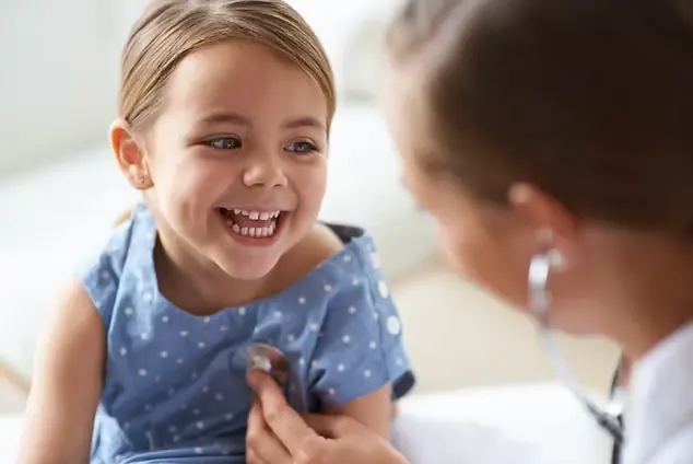Child in blue polka dot dress smiles as a doctor uses a stethoscope. Bright, cheerful setting with focus on child's joyful expression.