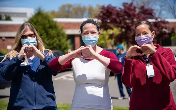 Three women wearing masks and colorful tops form heart shapes with hands outdoors on a sunny day, conveying gratitude and positivity.