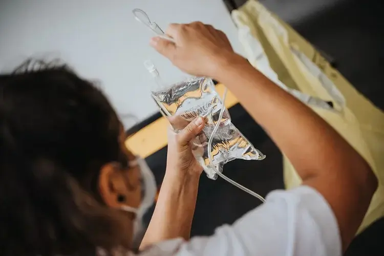 Person in white shirt adjusting a clear IV bag in a medical setting. Neutral tones, with yellow and white in the background.