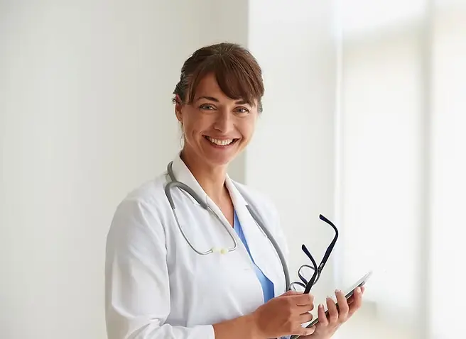 Smiling doctor in white coat with stethoscope holds phone and glasses, standing in bright room with blurred background.