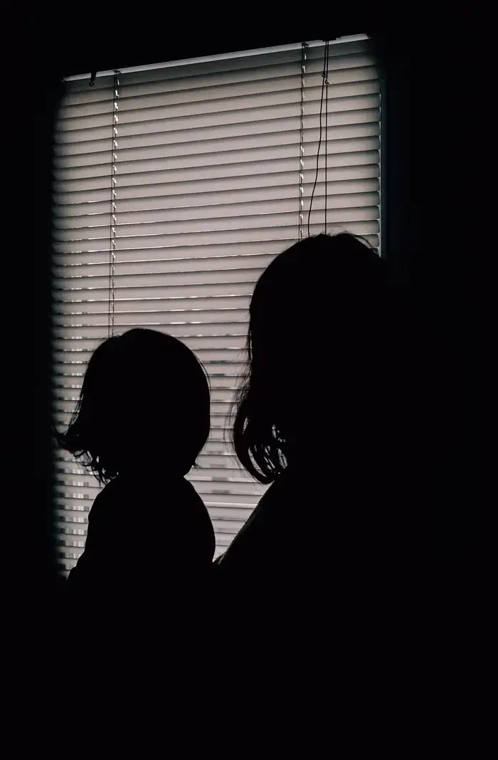 Silhouettes of a child and adult sit in front of a window with closed blinds. The ambient light casts a serene, contemplative mood.