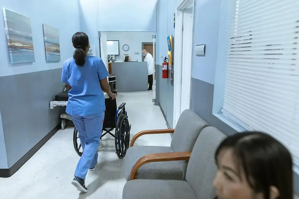 A nurse in blue scrubs pushing an empty wheelchair down a hospital corridor. 