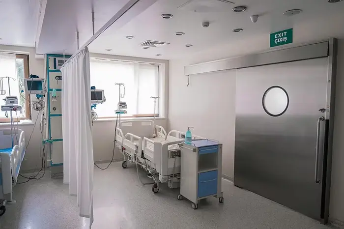 Hospital room with empty bed, medical equipment, and a closed metal door. Bright, sterile setting with "Exit" sign. Calm atmosphere.