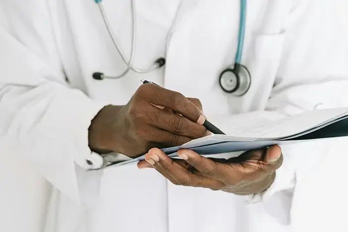 Doctor writing on a clipboard, wearing a white coat and stethoscope. Focus on hands. Neutral background, professional setting.