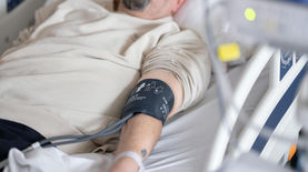 Elderly male patient lying in a hospital bed with a blood pressure cuff on his arm, representing inpatient monitoring and clinical observation.
