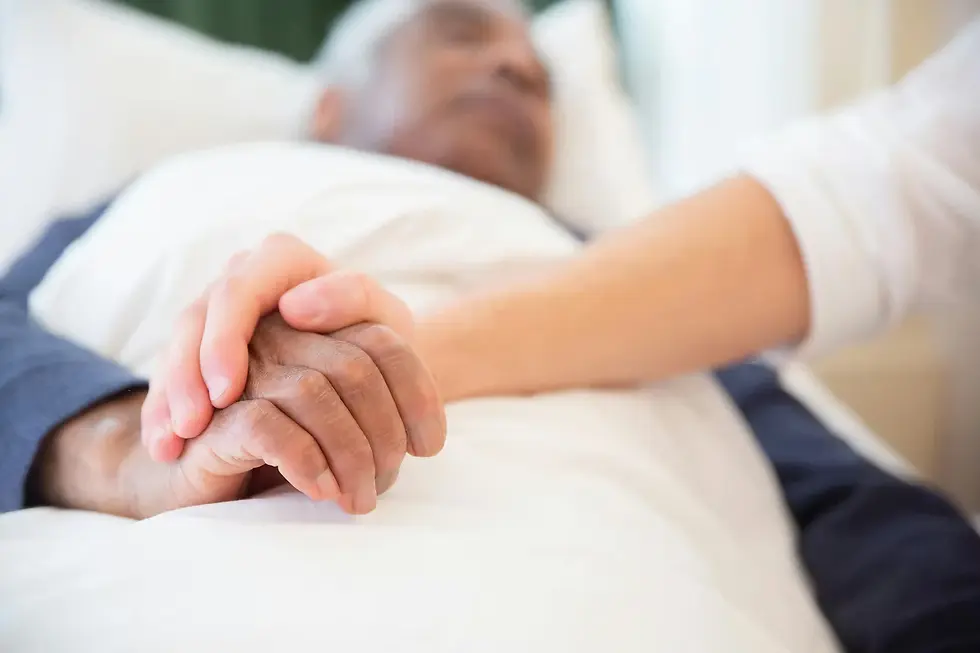 A person holds the hand of a resting individual in a hospital bed. The setting is calm, with white bedding and a supportive gesture.