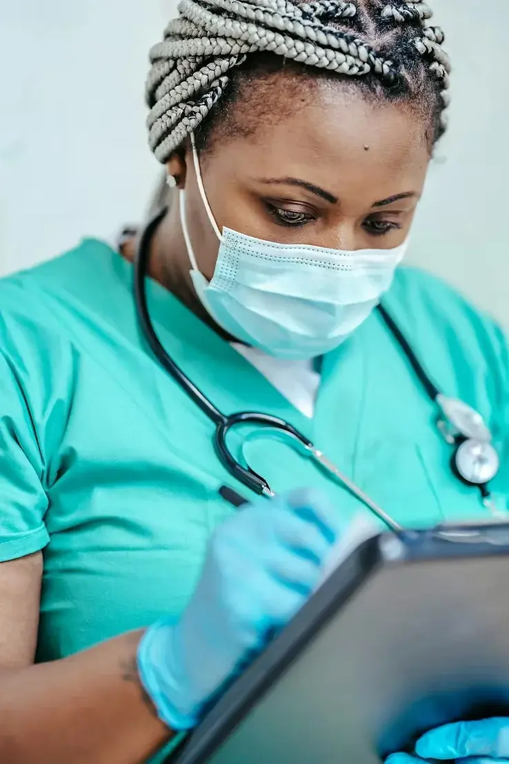 Nurse in teal scrubs, mask, and gloves writes on clipboard. Stethoscope around neck. Focused expression, neutral background.