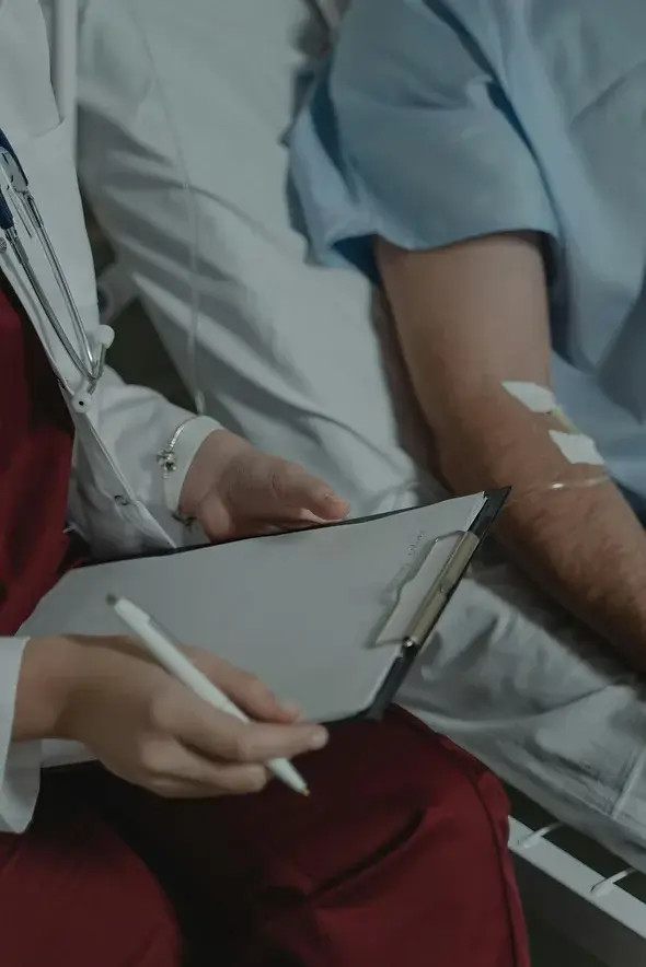A healthcare worker with a clipboard and pen sits beside a patient in a hospital bed. The patient has IV patches on their arm. Mood is clinical.