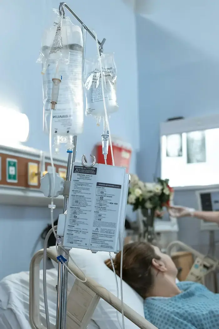Hospital room with a patient lying in bed, IV bags hanging beside them. Background shows medical equipment and flowers, creating a calm mood.