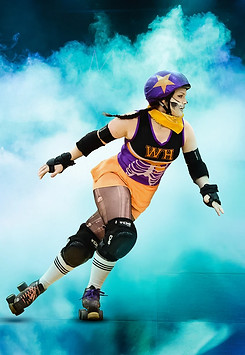 A photo of a skater doing a crossover. She wears orange and purple, a star on her helmet, ripped stockings, calf high socks, and bulky safety gear. The background is filled with blue fog.