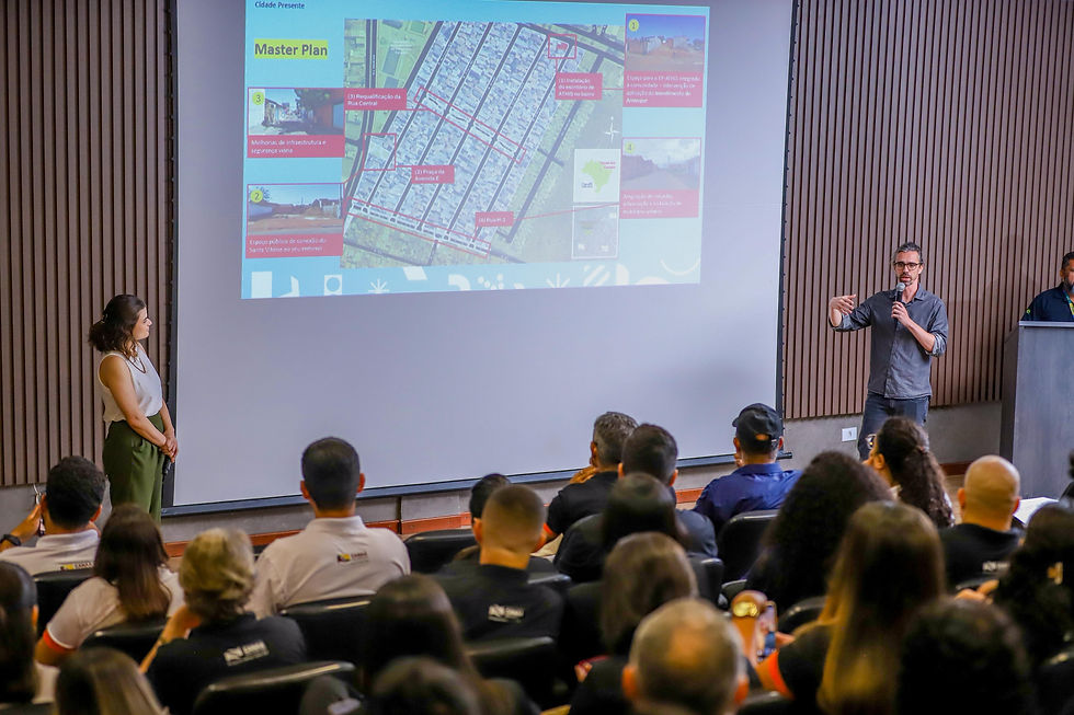 Fotografia de apresentação do Master Plan em auditório, com público atento e dois palestrantes à frente de um slide com mapa e informações do projeto.