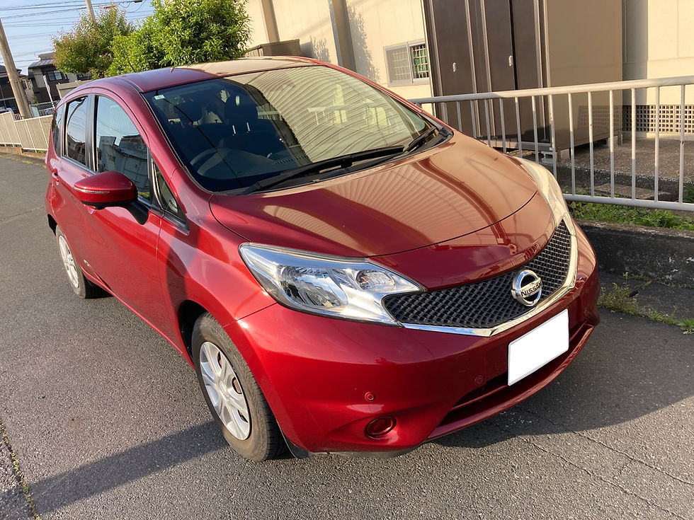 赤い日産のノートが住宅街の道路脇に駐車されている。背景には塀と植え込みが見える。晴れた日の午後の雰囲気。