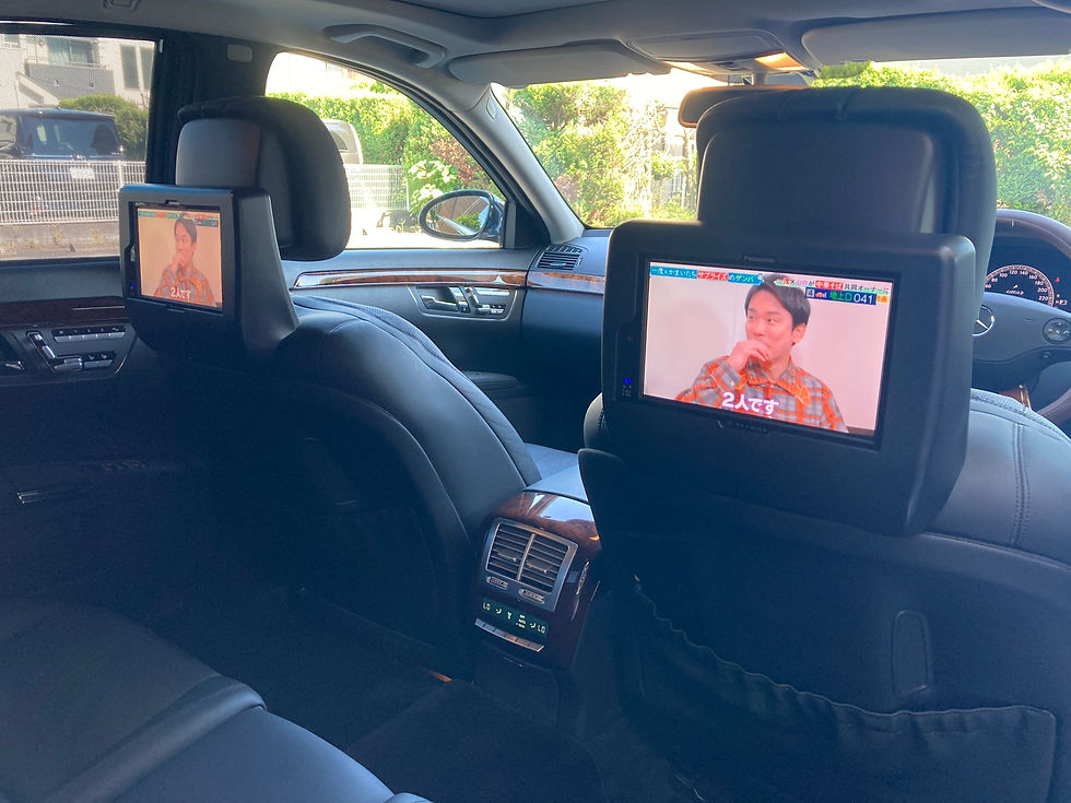 メルセデスベンツ後部座席の車内。前席ヘッドレストの2台のモニター。黒い革張りで日差しが差し込む。