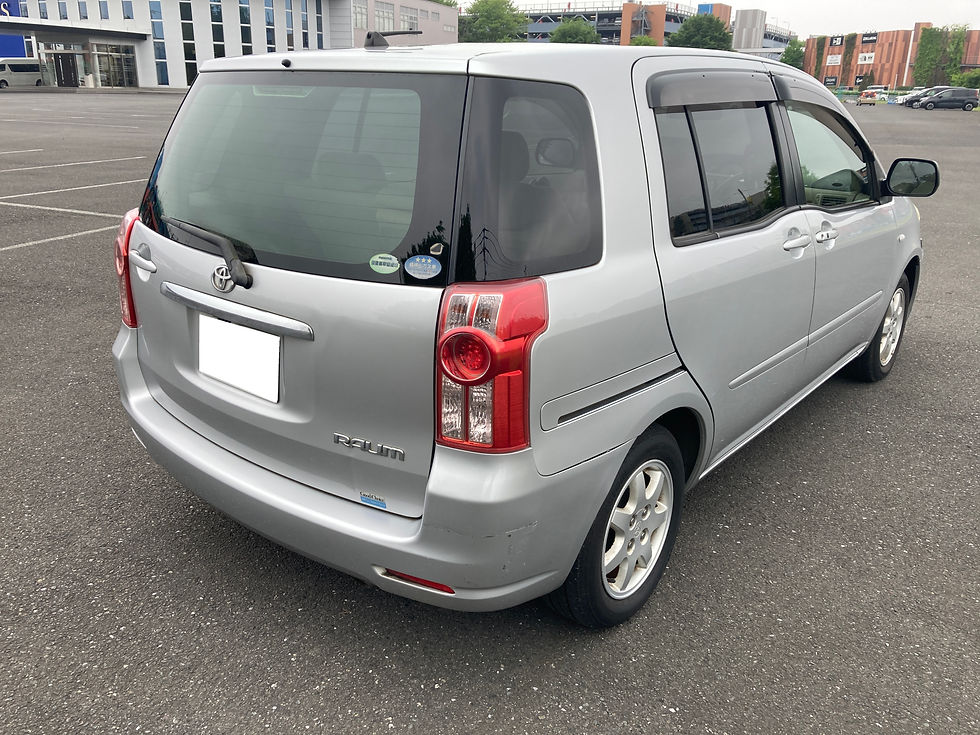 駐車場に停められた銀色のトヨタラウム、周囲に建物と木々。車のリアに小さいステッカー、静かな雰囲気。