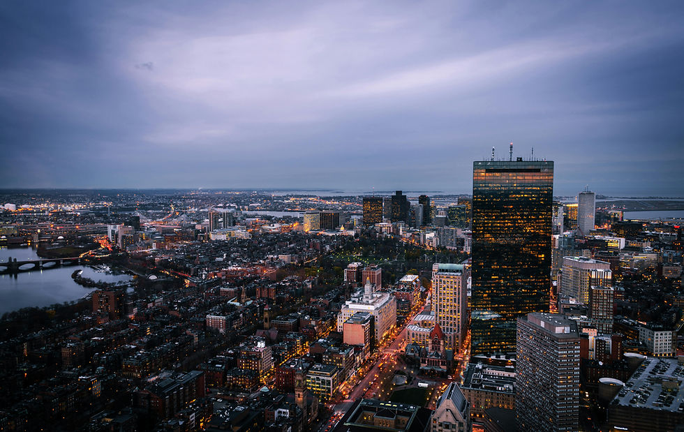 City skyline at dusk with illuminated buildings. A river and bridges are visible. Moody clouds and dim city lights create a calm atmosphere. Boston, Massachusetts