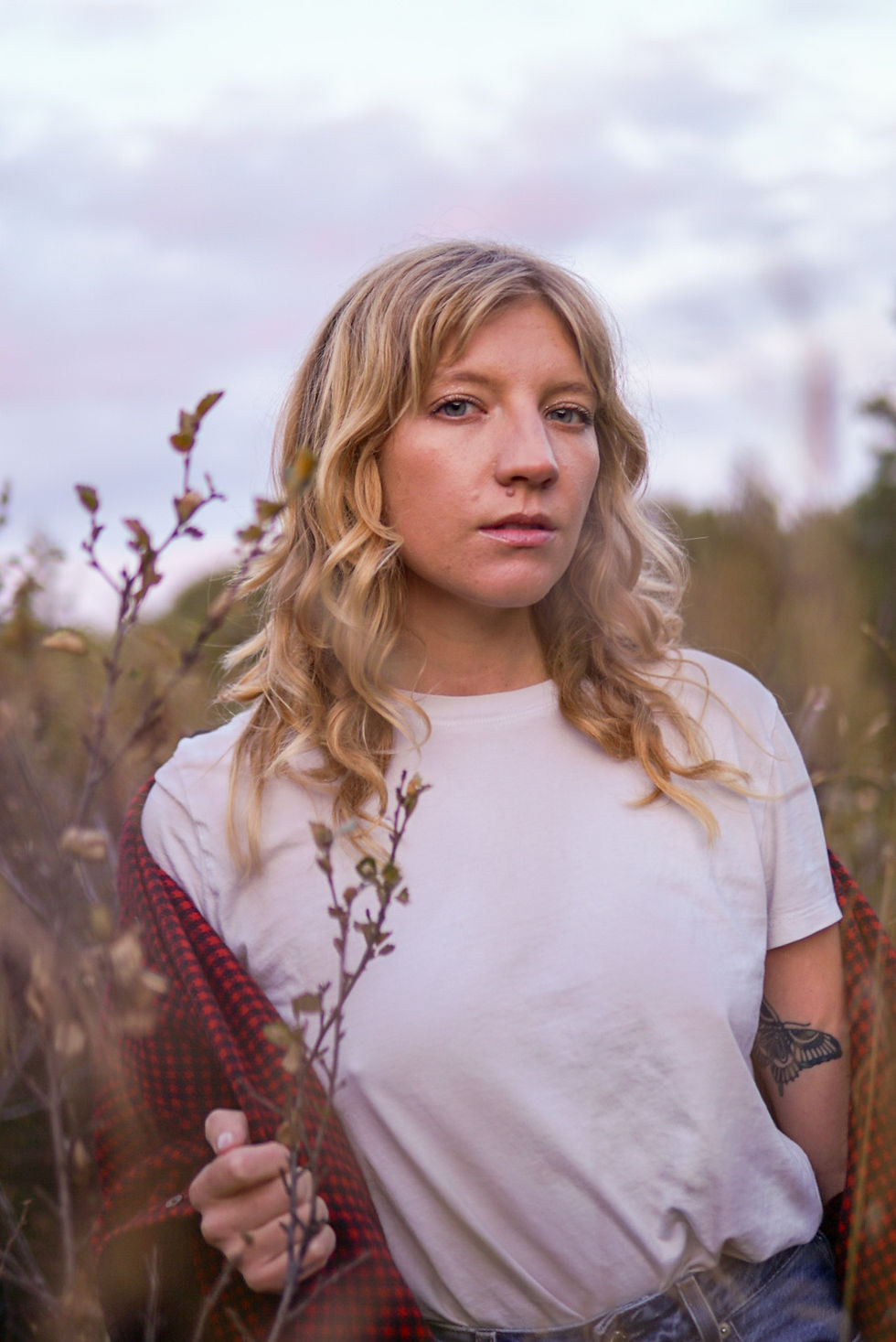 Clover Stieve stands in a field of tall grass under a soft, cloudy sky, wearing a white t-shirt with a red plaid layer draped over her shoulders, looking calmly at the camera with a natural, grounded expression.