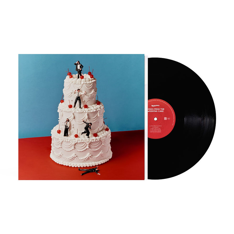 Blossoms' album Songs From the Wedding Cake on vinyl: the cover shows a three-tier white wedding cake with red cherries and tiny figures in black-and-white formal wear on its tiers, one lying face-up on the red surface in front, against a blue backdrop. A black record with a red label slides out of the sleeve at right.