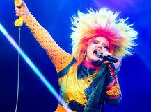 Caroline Kingsbury performing at Teragram Ballroom in Los Angeles, vocalist with a massive multicolored teased wig singing into a microphone while holding a yellow telephone receiver above her head in a rhinestone and sequined outfit under blue laser lighting