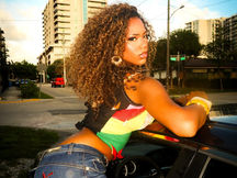 Sej leaning out of a black car on a city street at golden hour, wearing a colorful red, yellow, and green crop top with a black mesh overlay and low-rise jeans with “Bottoms” embroidered on the back pocket, with voluminous curly highlighted hair, a gold bracelet, and a city block of apartment buildings behind her